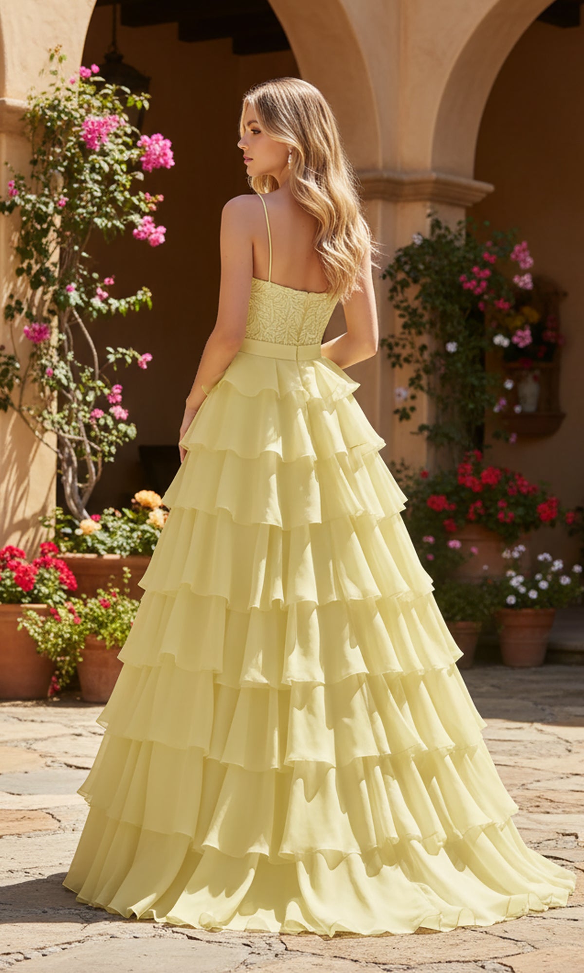A woman wearing the Long Prom Dress: PromGirl Exclusive 250202, a light yellow tiered ruffle gown, stands outdoors on a stone patio surrounded by potted plants and flowers.
